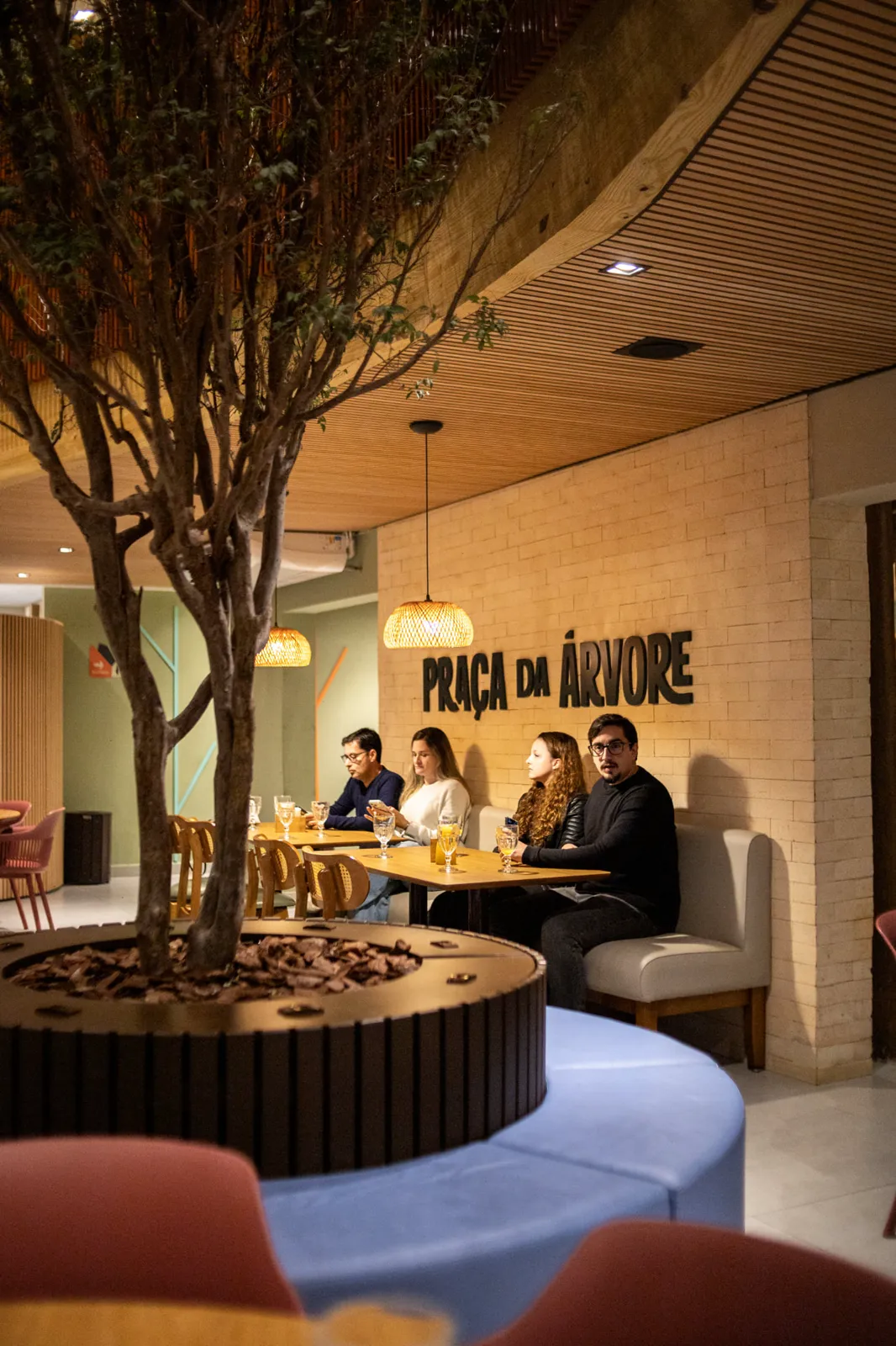 Salão com a árvore central, banco de madeira e o letreiro Praça da Árvore na parede