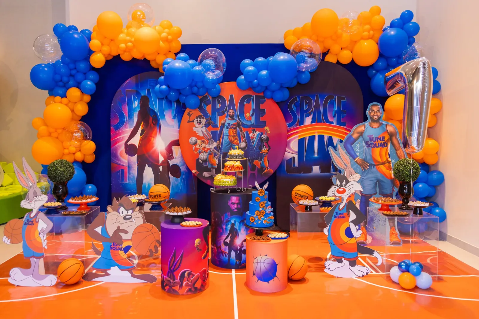 Mesa de festa com tema Space Jam: bolo azul, arcos de balões, bolas de basquete e painéis dos personagens